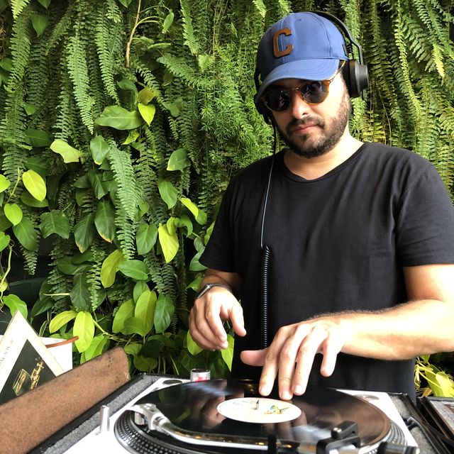 Leo Reis de boné azul, tocando vinil diante de um jardim vertical