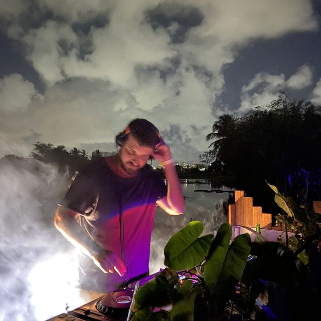 Leo Reis tocando ao ar livre, à noite, com nuvens e coqueiros ao fundo