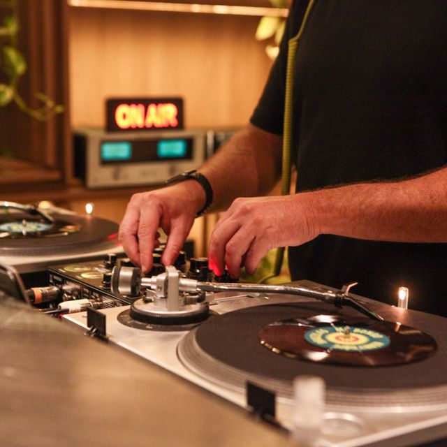 Mãos de Leo Reis no mixer e nos toca-discos, com a placa On Air ao fundo